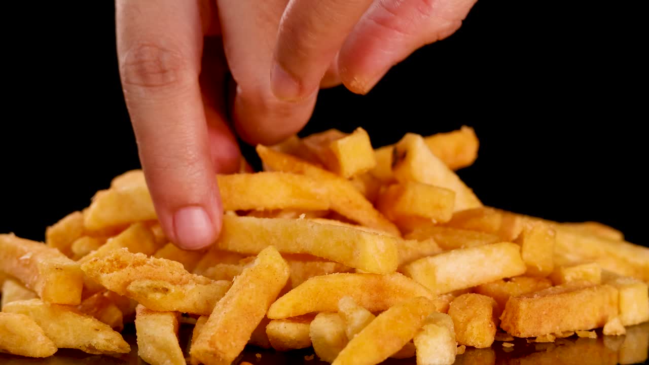 Human hand selects golden French fries from pile, close-up, studio lighting, black background, static shot