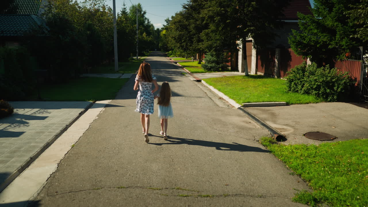 Back view of young woman running hand in hand with child along quiet suburban street, hair swaying in motion under bright sunlight with green lawns, trees, and gated houses on both sides
