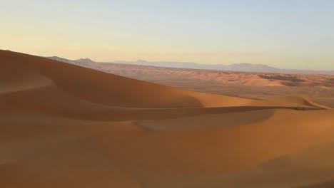 camera pans across a vast desert landscape, capturing the undulating dunes and distant mountain ranges bathed in the warm glow of the setting sun