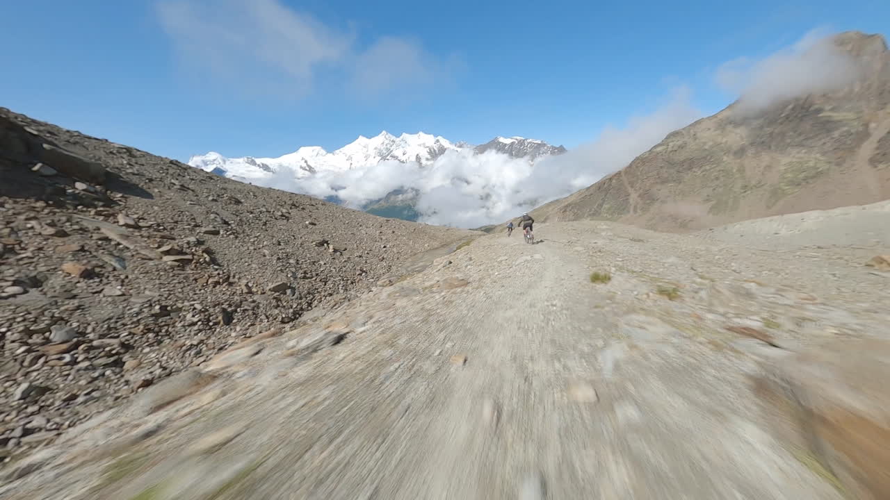 FPV drone follows two mountain bikers descending steep, exposed alpine trails above Saas-Grund and Saas-Fee on a clear bluebird summer day, showcasing epic terrain and speed