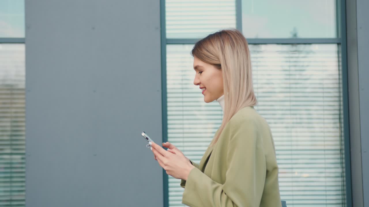 Side view of smiling young woman looking at mobile phone walking in the street to the office beside green trees pine. Outdoors