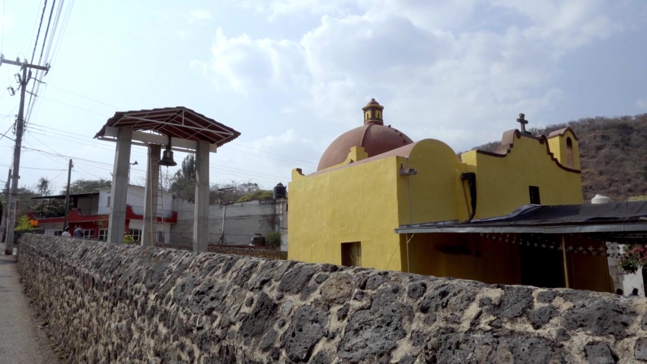 Piccola chiesa gialla con cupola e campanile dietro un muro di pietra, ambientazione rurale con colline e cielo, architettura tradizionale catturata alla luce del giorno con dintorni pacifici.