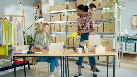 diseñadora afroamericana empacando ropa ordenada en cajas mientras un diseñador le muestra una pieza de tela en una tienda de ropa de moda