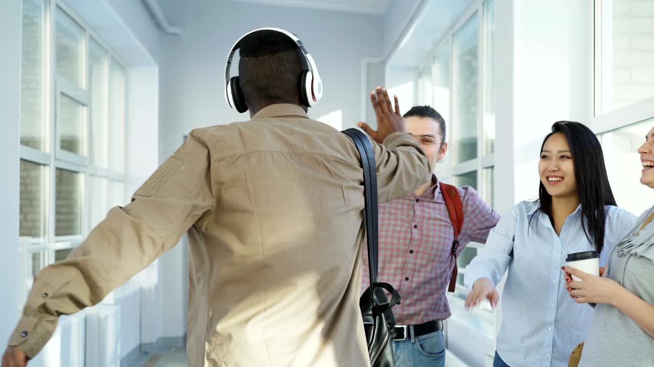 Toma con dolly de un joven y apuesto estudiante afroamericano caminando y bailando en un pasillo blanco de la universidad, notando a sus amigos, saludándolos y continuando escuchando música con auriculares grandes