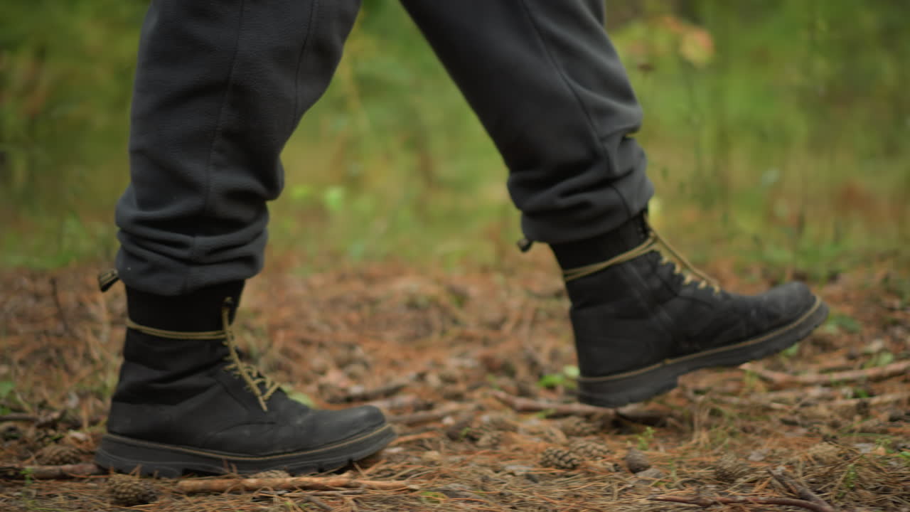 bota de senderismo en ángulo bajo cruzando un sendero cubierto de musgo, pasos rítmicos y firmes bajo el dosel de pinos, cordones amarillos y botas oscuras, pantalones de chándal grises rozando la corteza y la hojarasca, enérgica caminata matutina a través del verde