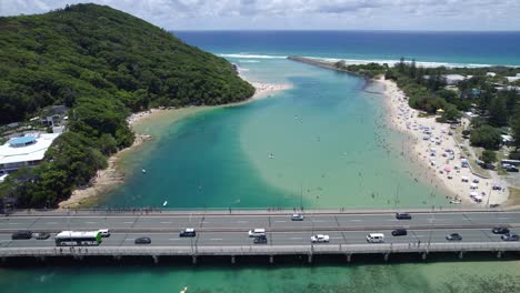 Vehículos-Circulando-Por-El-Puente-Tallebudgera-Creek-Con-Un-Paisaje-De-Playa-Llena-De-Gente-Durante-El-Verano-En-Queensland,-Australia