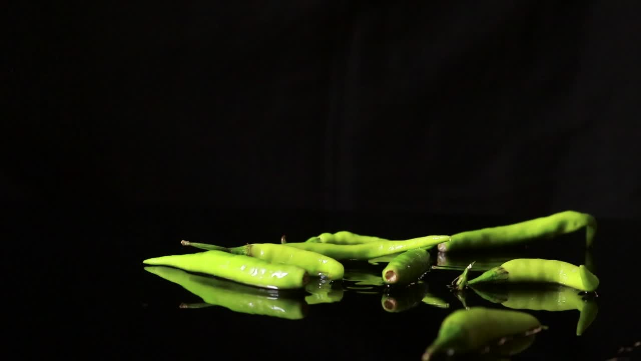슬로모션 그린 버드아이 칠리 페퍼는 각각의 뜨겁고 매운 신선한 요리 야채에 반짝이는 물웅덩이가 있는 흰색 접시 표면에 떨어집니다.
