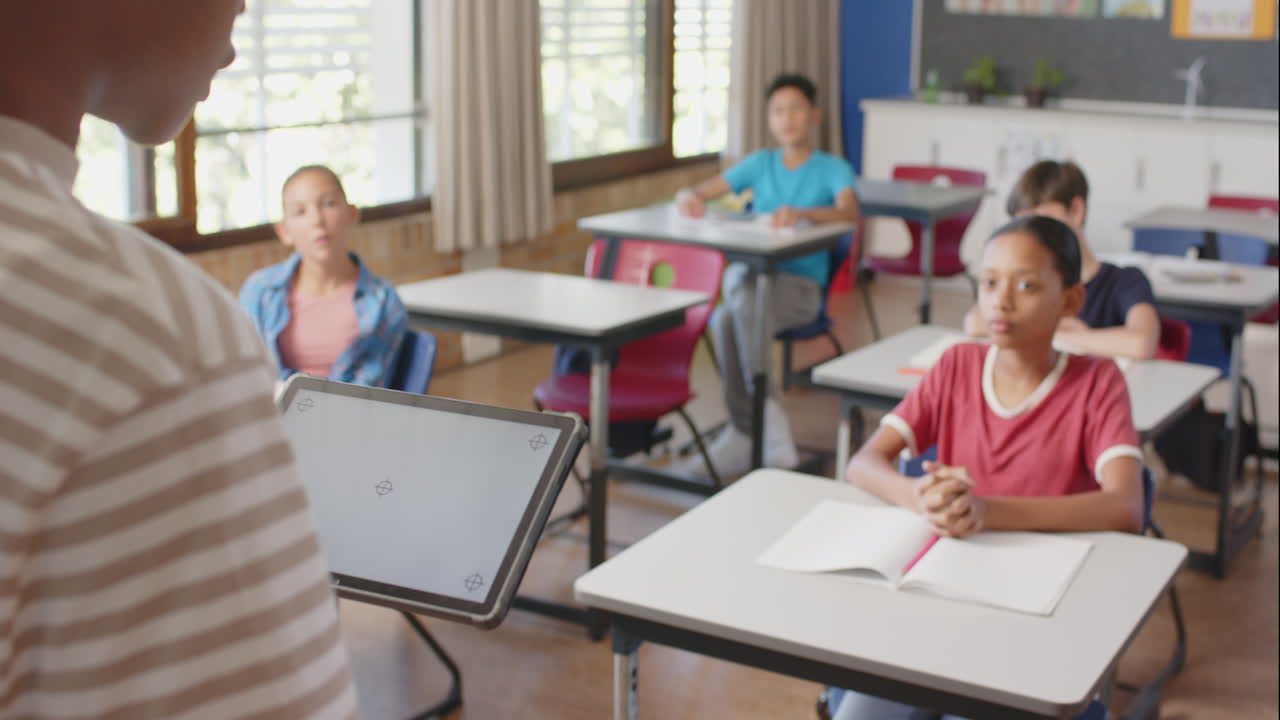 Ve škole učitel používá tablet, zatímco studenti pozorně poslouchají ve třídě.