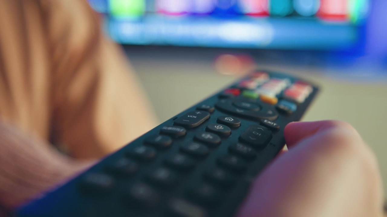 Low-angle close-up shows hand holding textured plastic remote control while thumb presses top buttons and device tilts toward blurred glowing screen, suggesting channel change, menu navigation, and