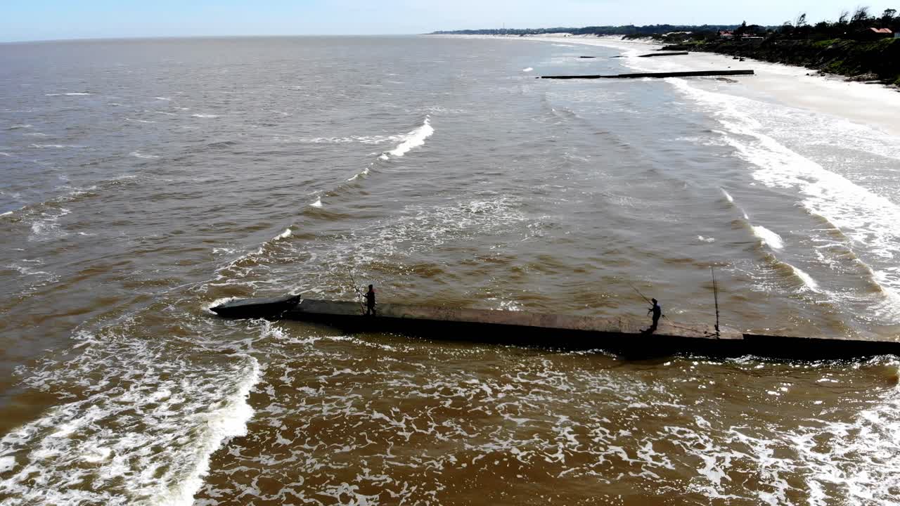video aereo de la playa bajando al espigón con dos hombres pescando un dia con olas