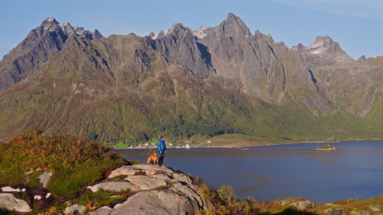 A man and dog stand on a rocky hill overlooking Vestarelen mountains and lake
