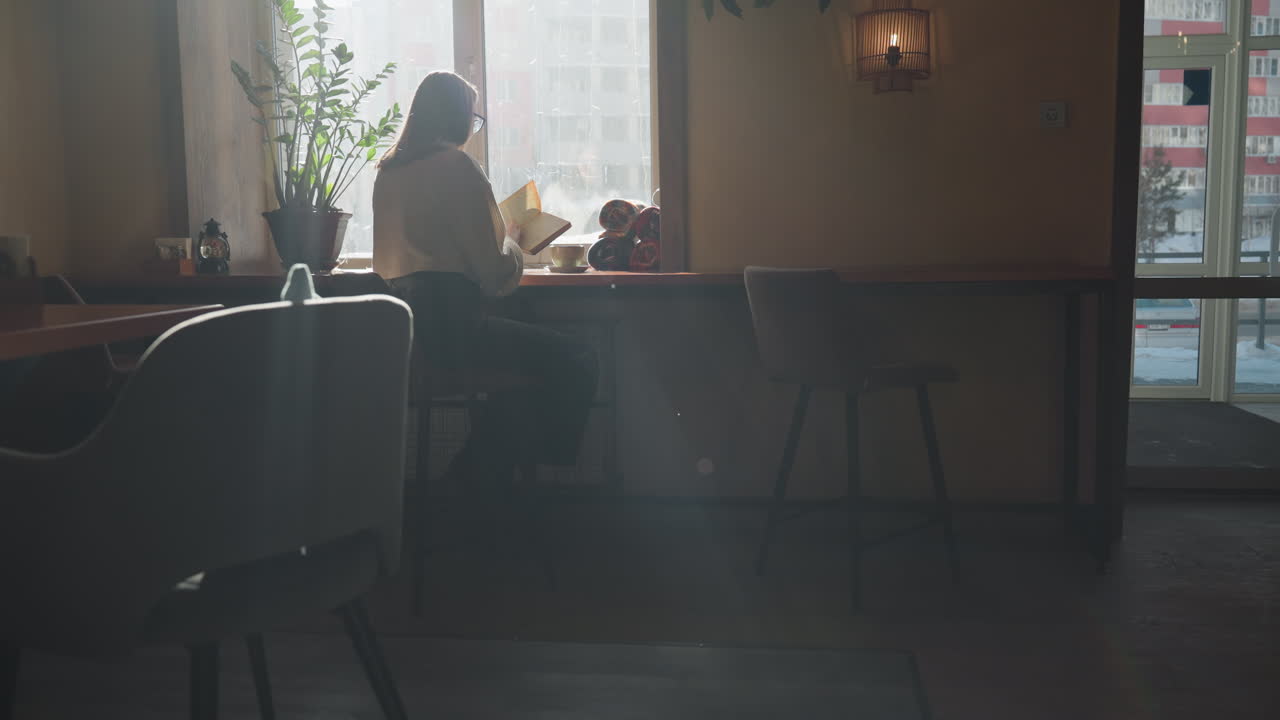 Young adult woman flipping through book near sunlit window in cozy interior, warm rays highlighting her white sweater as steam rises from coffee cup beside her, surrounded by plants and calm ambiance