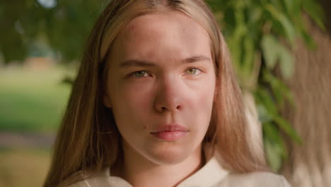 close-up of young lady with intense expression, sunlight reflecting softly on her face, adding warmth and depth to her look, with blurred green foliage in background