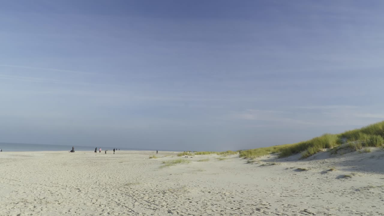 A sand dune with tufts of grass intermingles with a wide sandy beach where a few tourists are relaxing