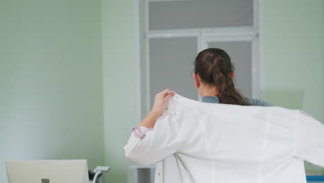 medical practitioner wears uniform in bright clinical room then walks toward desktop computer to begin daily work routine seated on chair while adjusting posture with focus and professionalism