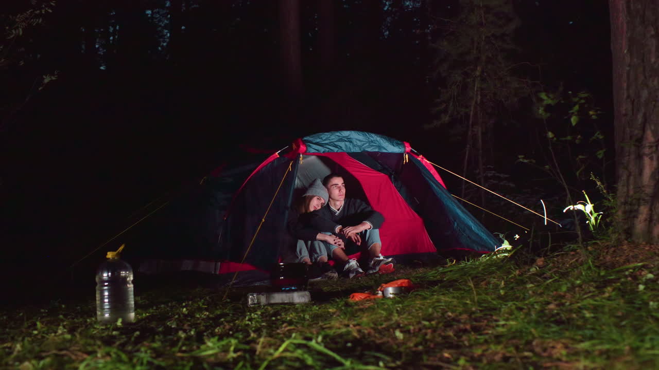 Lady rests head on boyfriend shoulder as couple sits quietly inside tent surrounded by forest at night, with camping gear and warm light capturing peaceful outdoor moment