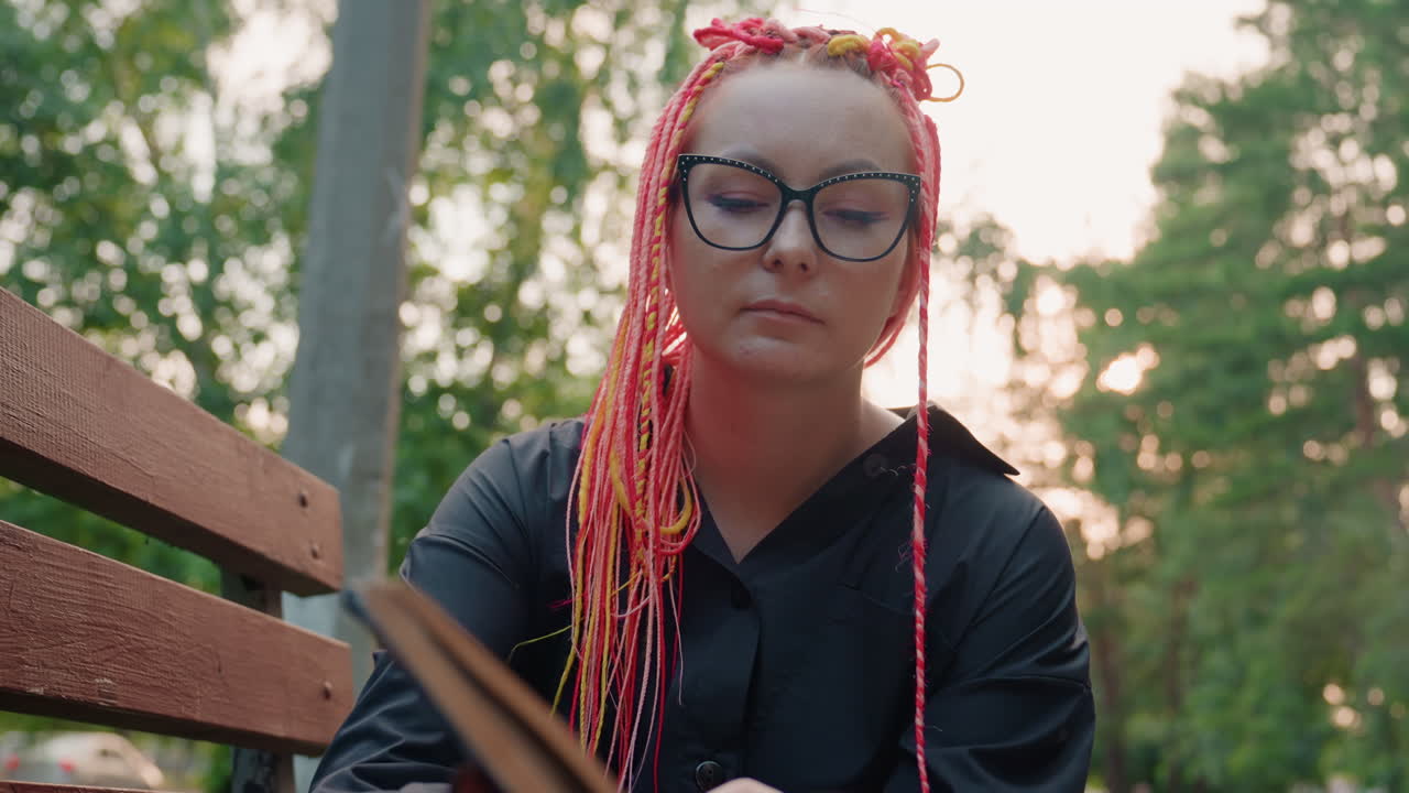 lady reading outdoors, girl with colorful dreadlocks focused on reading in quiet park setting, woman sporting pink dreadlocks quietly immersed in reading in tranquil outdoor environment