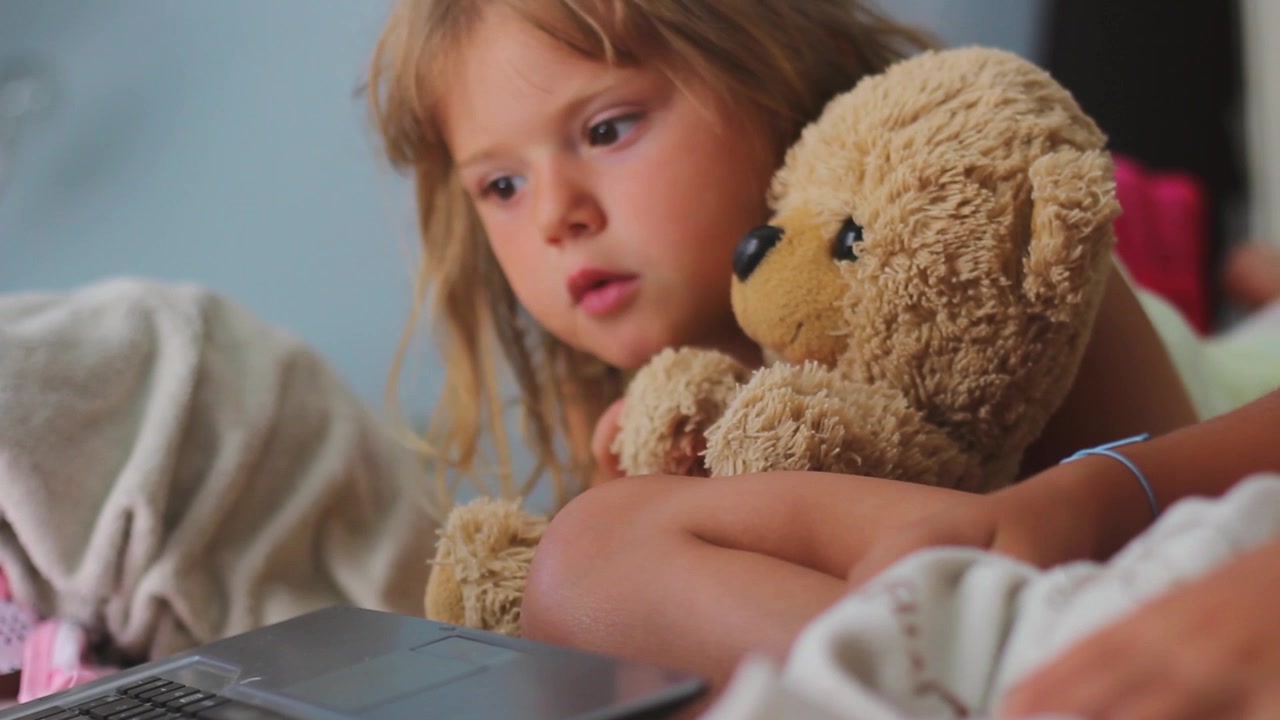 Primo piano di una bambina sdraiata sul letto con la sorella, mentre tiene il suo orsetto di peluche e guarda video sul laptop
