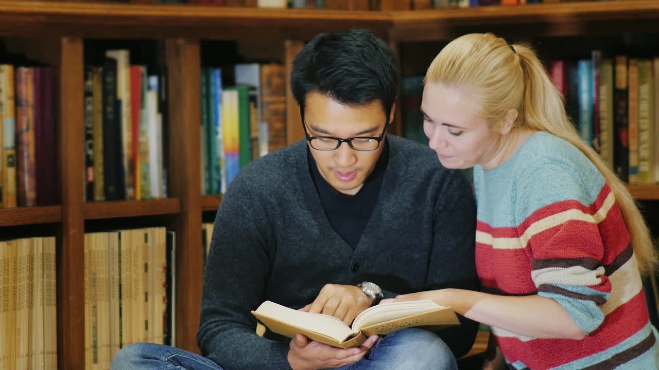 Asiatisk mann med briller og kaukasiske kvinner står sammen og ser på boken på biblioteket.