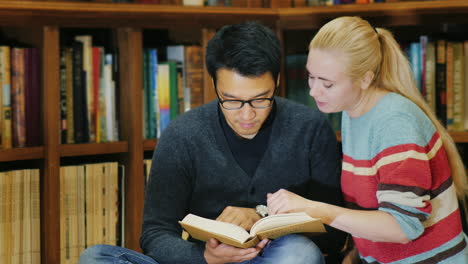 Hombre-Asiático-Con-Gafas-Y-Mujeres-Caucásicas-De-Pie-Juntos-Mirando-El-Libro-En-La-Biblioteca