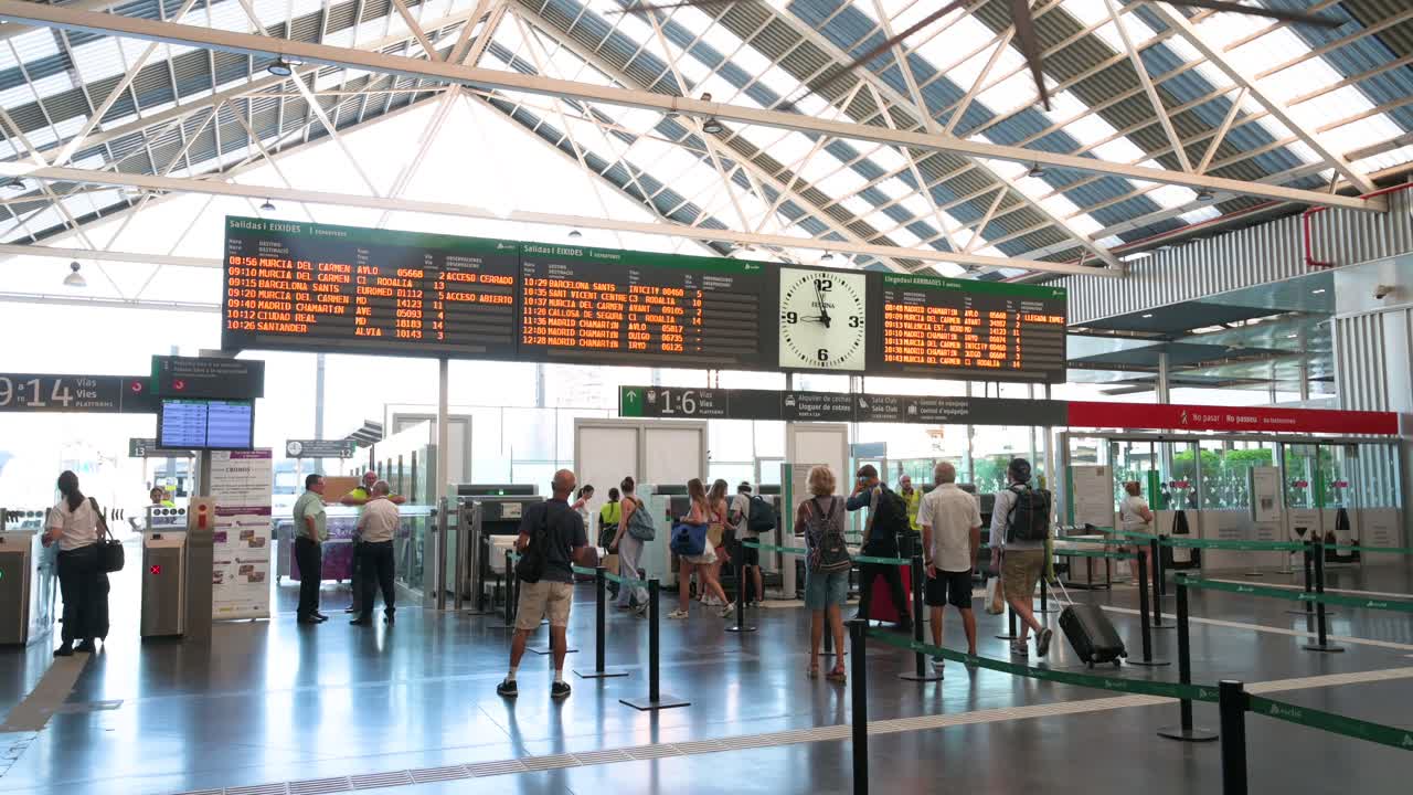 Wide shot of passengers arriving at the main terminal hall of Alicante train station and undergoing luggage security checks in Alicante, Spain.
