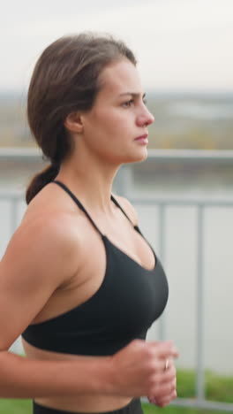 a focused woman in black jogs by an iron fence near a river, showcasing a determined expression as she runs in the park. in the background, a bridge and flowing river enhance the serene environment