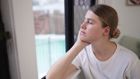 Joven-Mujer-Caucásica-Sentada-Y-Mirando-Por-La-Ventana-Reflectante-En-Casa,-4k