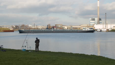 fisherman on shore preparing rod while a cargo ship moves over river