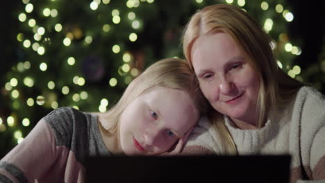 mom and daughter are looking at the laptop screen together. sitting in front of the blurred lights of the christmas tree