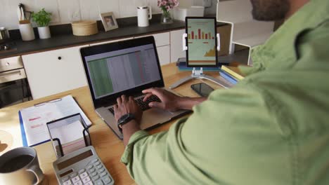 african american man sitting at table in kitchen, using laptop and tablet