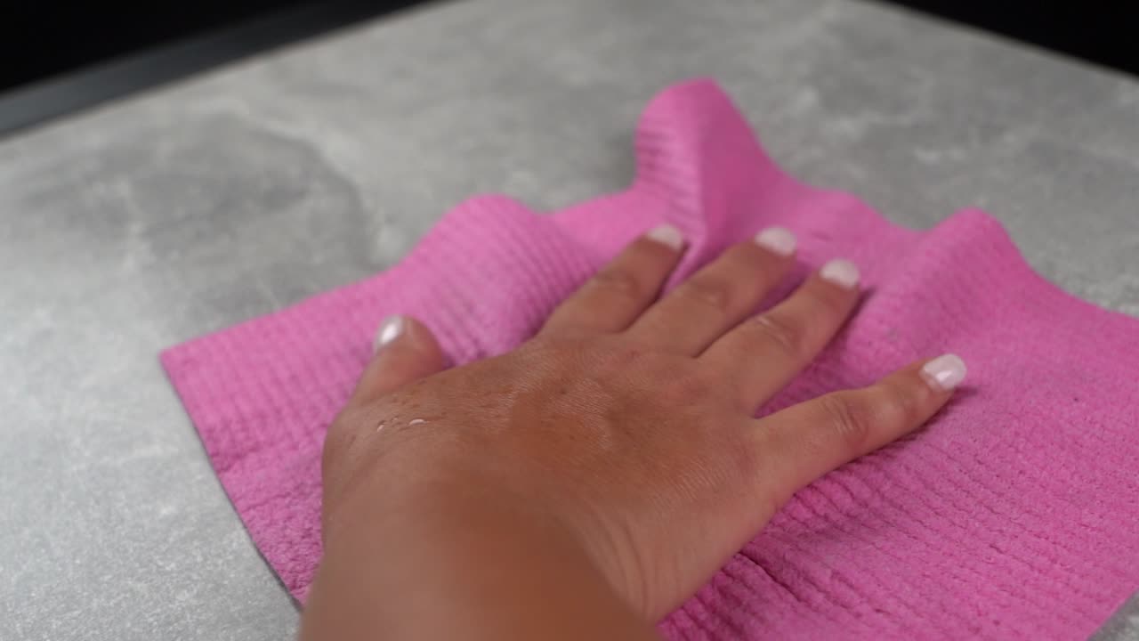 женщина с окрашенными ногтями, использующая розовую ткань для чистки серого стола