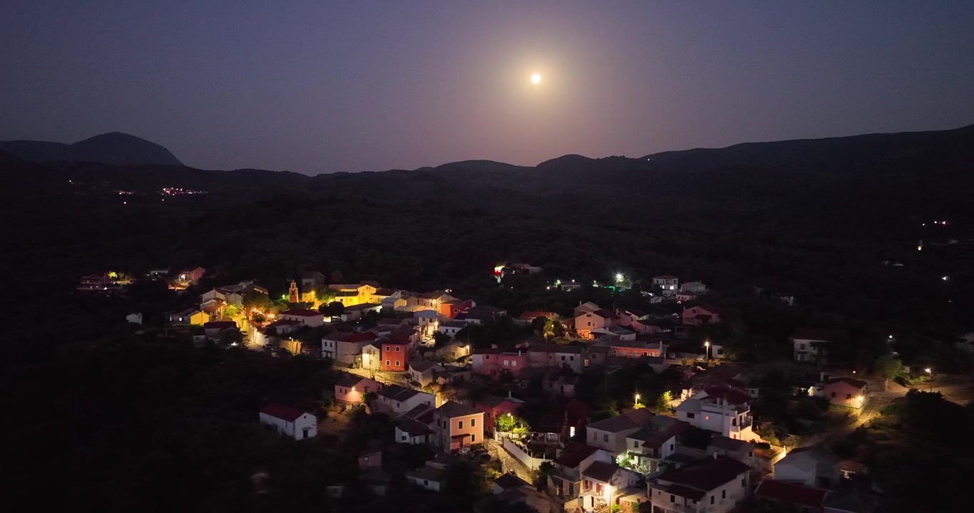 Tomada aérea de drone das luzes da vila de Agioi Douloi à noite sob uma lua cheia brilhante, Corfu, Grécia