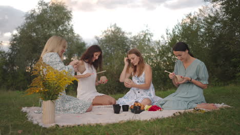 a group of young beautiful women concentrates on drawing patterns on clay products with the help of tools decorate molded objects talking smiling in a meadow in nature in an open space.