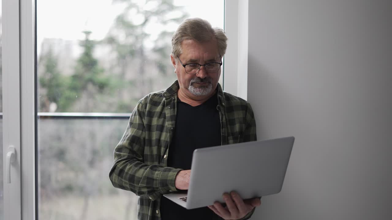 Senior older mature man typing on laptop browsing internet standing next to the window