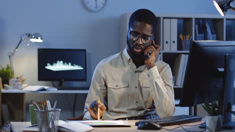 young worried businessman talking on the phone and consulting about some documents of project sitting at desk in the office at night