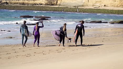 Toma-Mediana-De-Surfistas-Caminando-Con-Sus-Tablas-De-Surf-Y-Trajes-De-Surf-Sobre-Una-Hermosa-Playa-De-Arena-Durante-Las-Tranquilas-Olas-Del-Mar