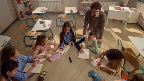 Students-sitting-at-round-table-in-classroom.-Teacher-explaining-task-of-lesson