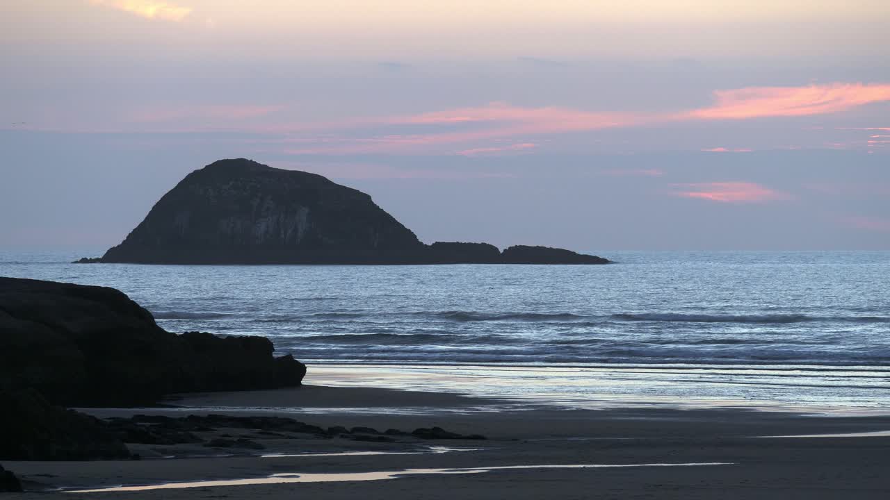 océano tranquilo con silueta de montaña en el fondo durante la puesta de sol en la playa de muriwai, auckland, nueva zelanda