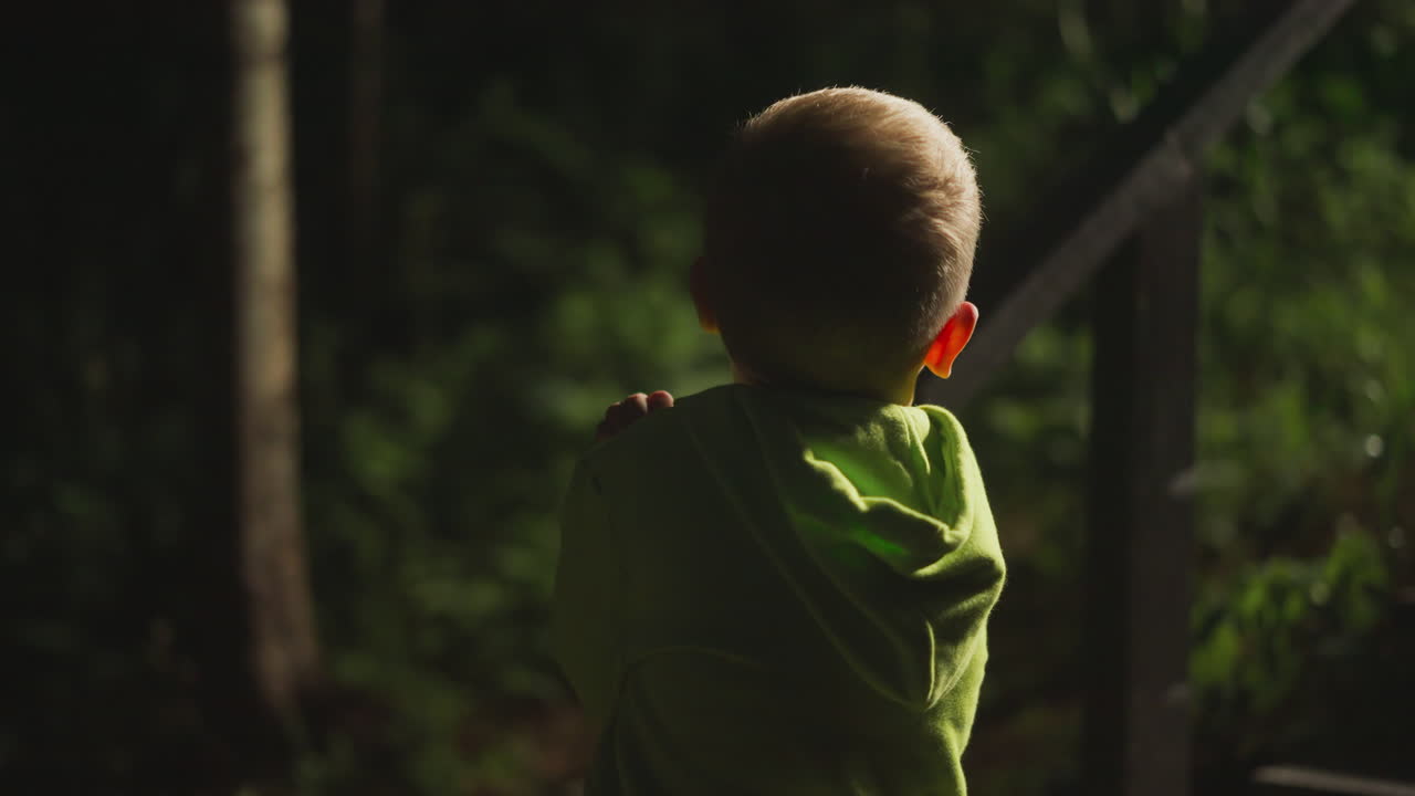 Little boy in bright light green hoodie stands under lantern near dark evening forest. Child spends time in nature playing and relaxing against of wooden stairs