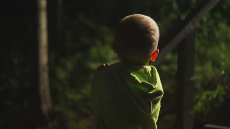 little boy in bright light green hoodie stands under lantern near dark evening forest. child spends time in nature playing and relaxing against of wooden stairs