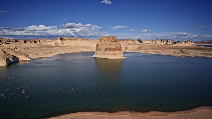 Drone flies over the deep blue waters of Lake Powell, spotlighting a dramatic rock formation against a rugged canyon landscape.
