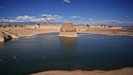 drone flies over the deep blue waters of lake powell, spotlighting a dramatic rock formation against a rugged canyon landscape.