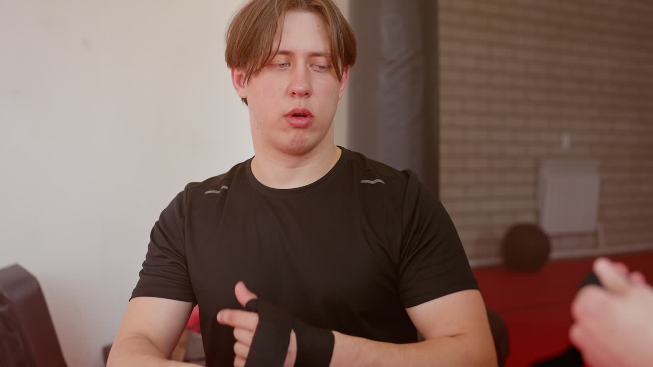 Athlete in black sportswear prepares hand wrap before training on red mat in gym, showing focus, strength, and discipline during pre-fight routine, emphasizing preparation, combat sport dedication