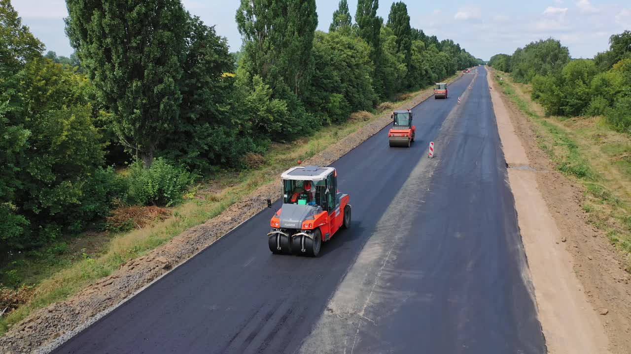 Road rollers working on the highway. Trucks leveling up fresh asphalt on the road among green nature. Asphalt road construction. Aerial view. Motion camera back.