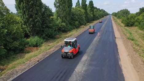 road rollers working on the highway. trucks leveling up fresh asphalt on the road among green nature. asphalt road construction. aerial view. motion camera back.