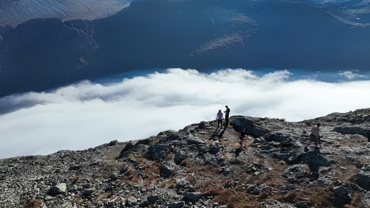 Drone circles above three people and a dog moving away from a cliff edge, revealing Naeroyfjord with clouds below