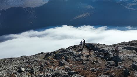 drone circles above three people and a dog moving away from a cliff edge, revealing naeroyfjord with clouds below