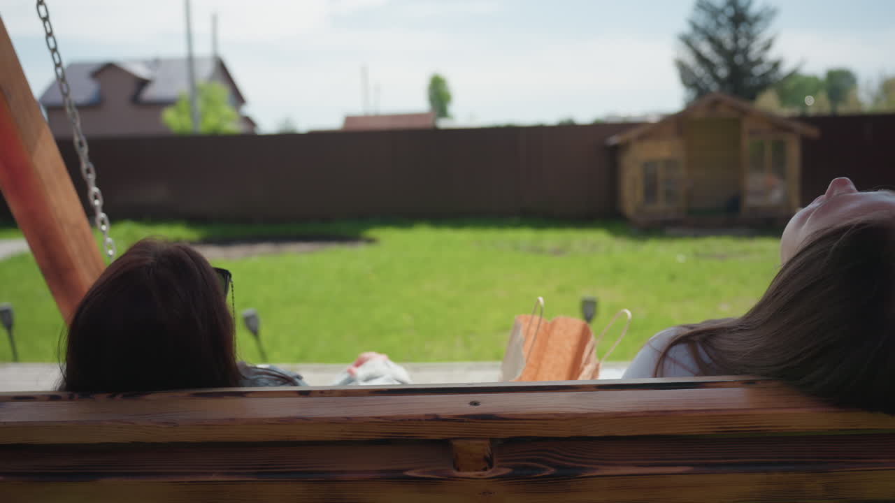 Rear view of two women gently swinging on wooden bench in sunny backyard, one wears sunglasses, other leans back with shopping bag beside her. Green grass, wooden fence, and clear sky