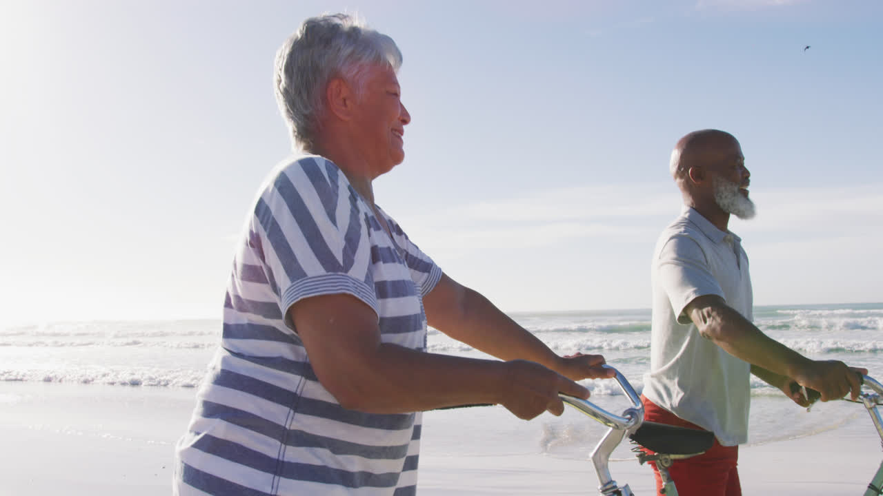 Smiling senior african american couple walking with bicycles at the beach