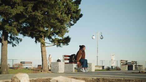 a girl wearing a brown coat, blue jeans, and white shoes sits alone on a bench in an urban park, her hand resting on the bench beside her black backpack. she looks disconsolate and lost in thought